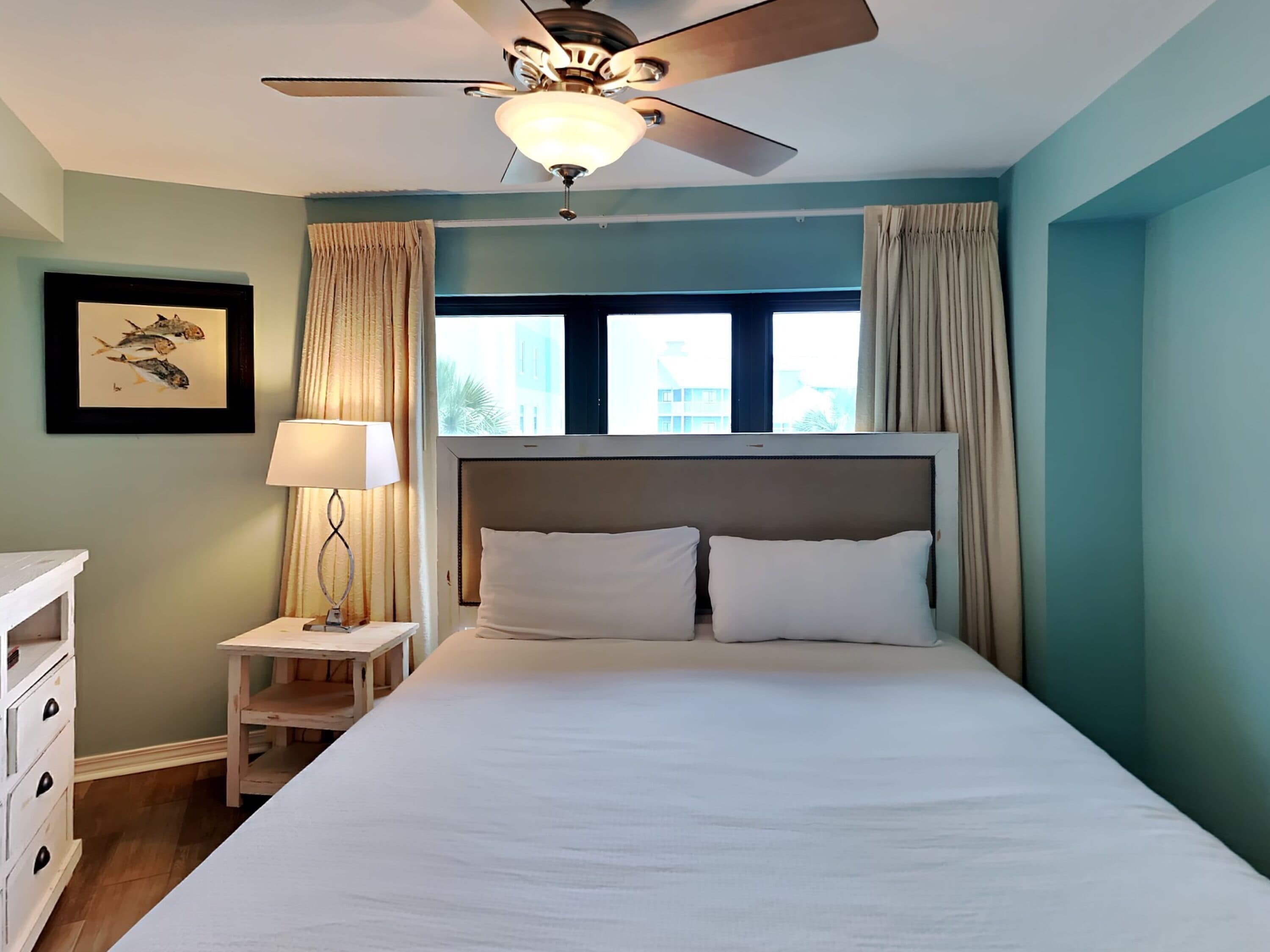 A bedroom with a large bed, a ceiling fan, a nightstand with a lamp, a framed picture on the wall, and a window with curtains.