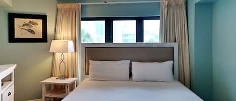 A bedroom with a large bed, a ceiling fan, a nightstand with a lamp, a framed picture on the wall, and a window with curtains.