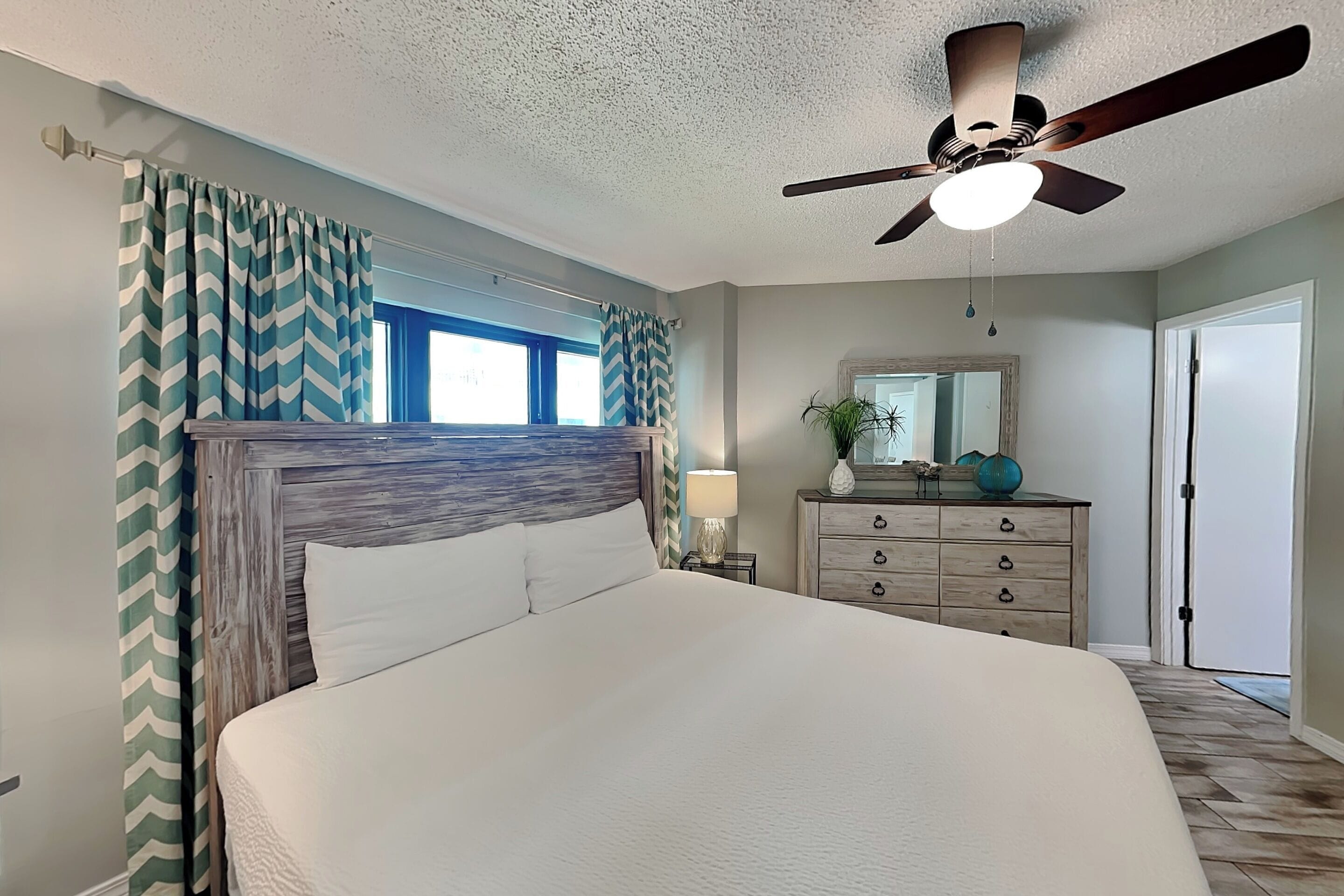 A bedroom with a bed, a ceiling fan, a dresser, a mirror, and a window.