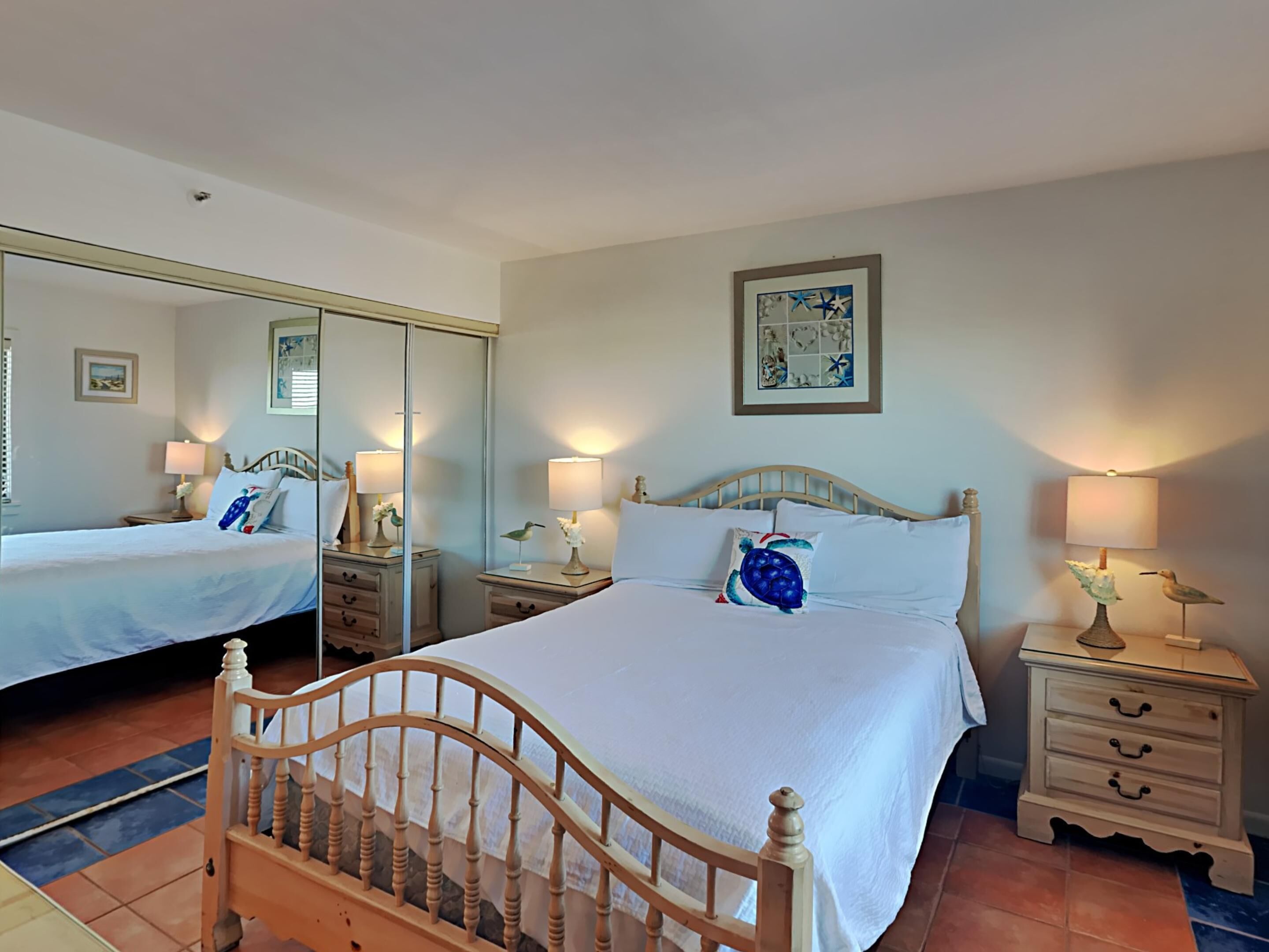 Ein Hotelzimmer mit einem großen Bett, zwei Nachttischen, einem Spiegel und einem an der Wand hängenden Kunstwerk.