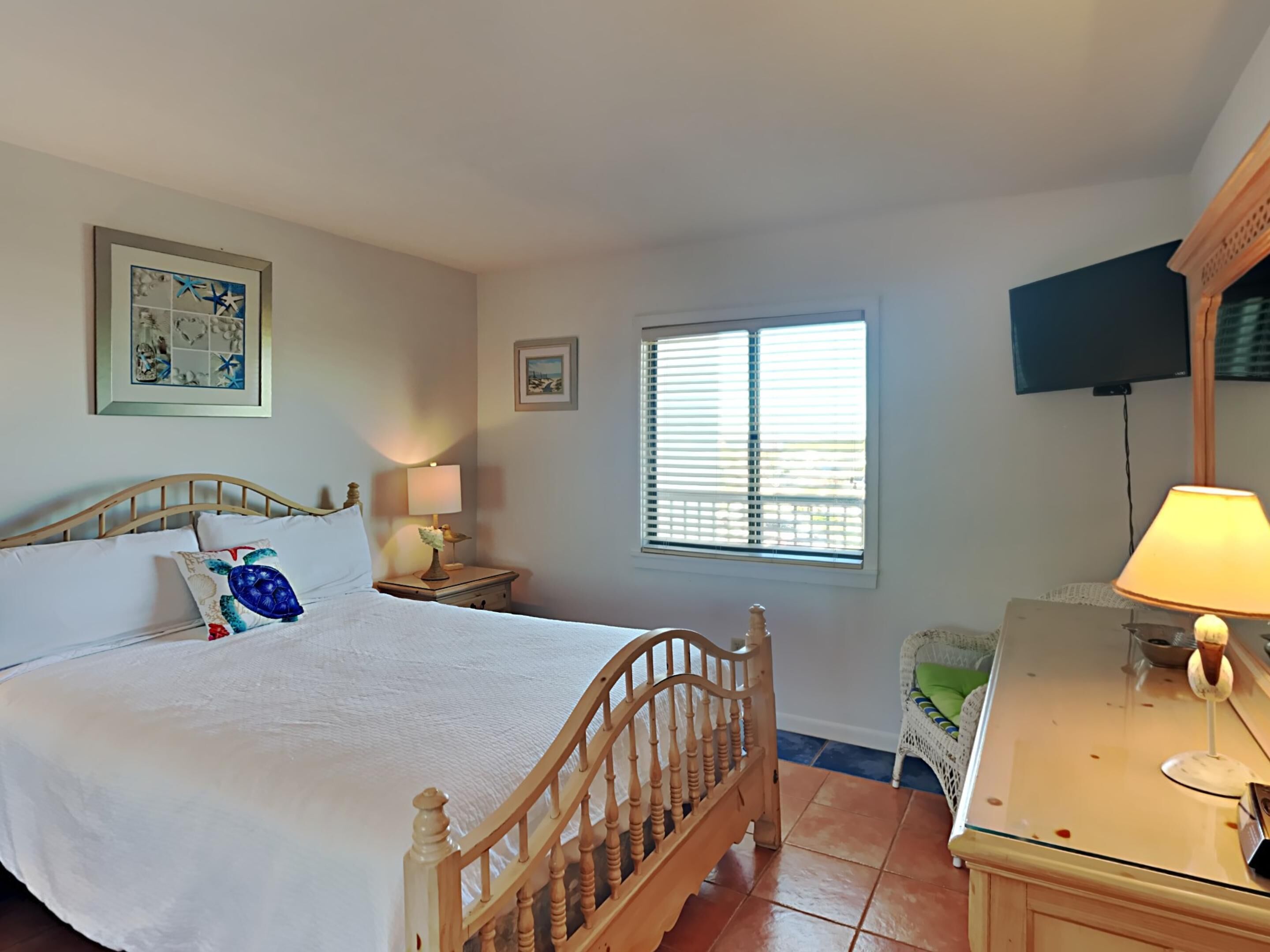 Une chambre avec un lit en bois, une table de chevet avec une lampe, un téléviseur fixé au mur et une fenêtre avec des stores.