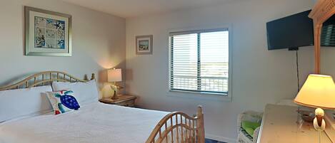 Une chambre avec un lit en bois, une table de chevet avec une lampe, un téléviseur fixé au mur et une fenêtre avec des stores.