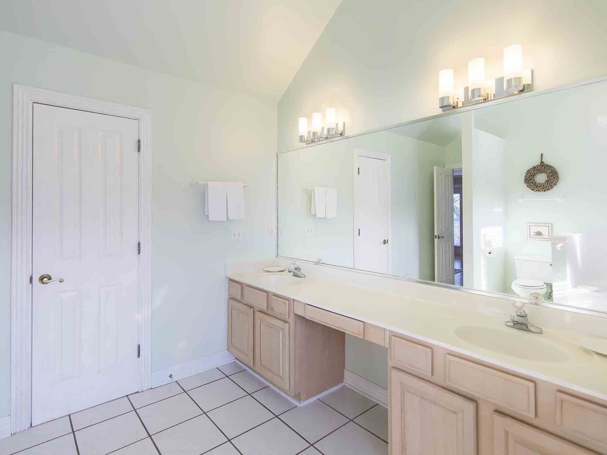 A bathroom with a large mirror, two sinks, and a toilet. There are white towels hanging on the wall and a door with a doorknob.