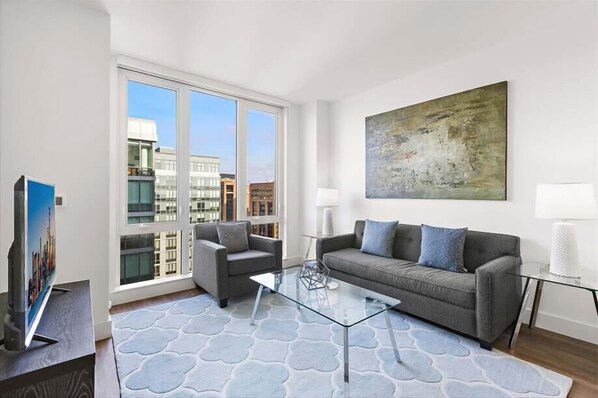 Ein modernes Wohnzimmer mit einem grauen Sofa, einem Glastisch und Blick auf die Stadt durch große Fenster.