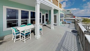 Et kysthus med en overdækket terrasse, store vinduer og en træterrasse med udsigt over stranden.