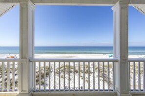A beach view from a covered porch with white railings, sandy beach, and ocean. - Simply The Best by Resort Collection (Panama City Beach)