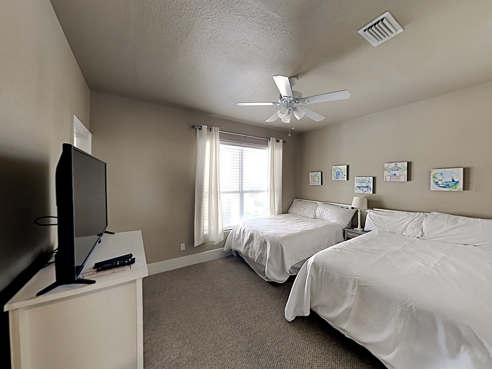 A hotel room with two beds, a ceiling fan, a TV, and artwork on the walls.