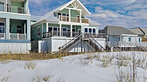 En ejendom ved stranden med et toetagers hus, en overdækket veranda og en trappe op til en terrasse.