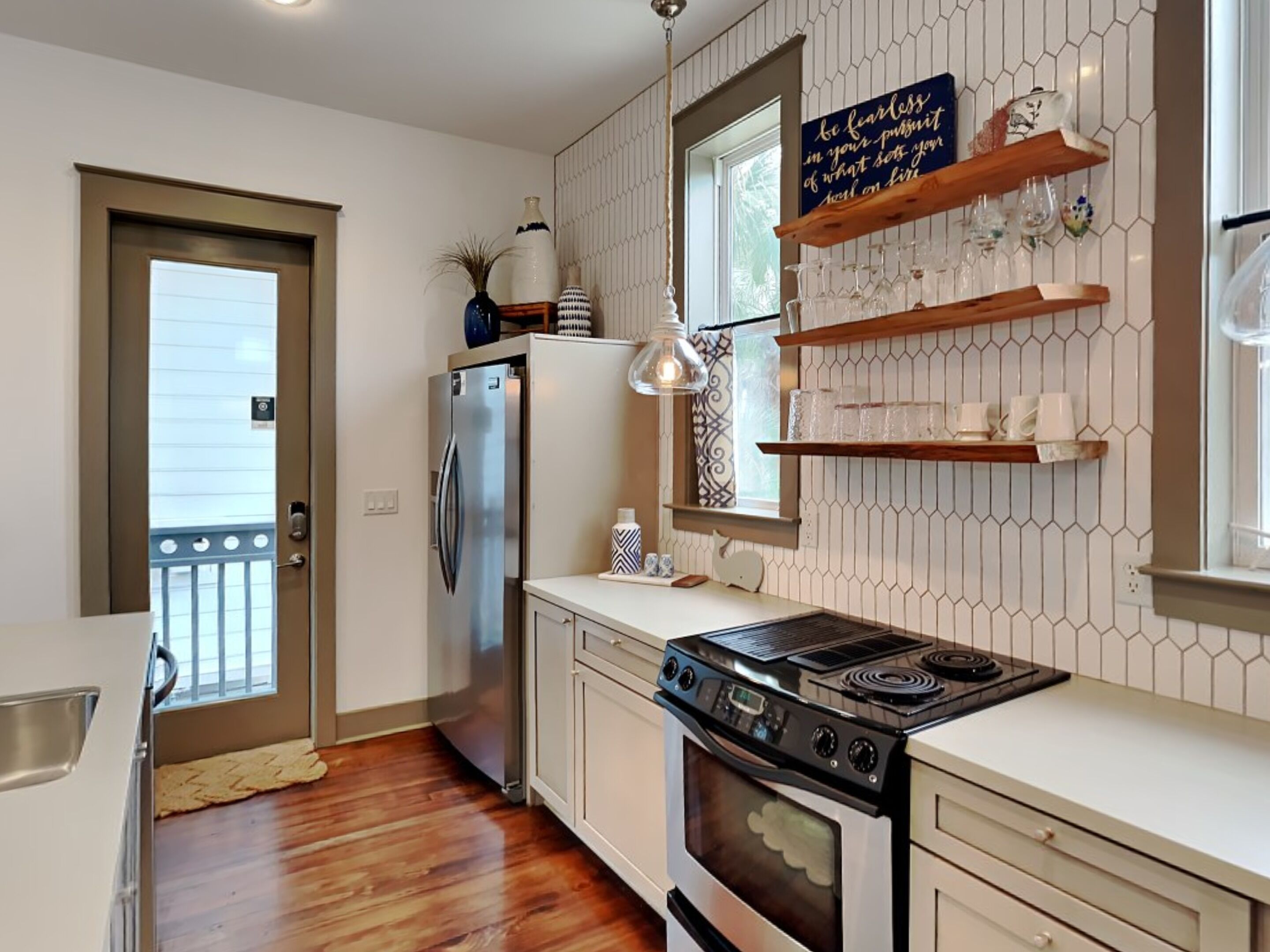 Una cocina moderna con un refrigerador de acero inoxidable, un horno y una estufa integrada. Hay repisas de madera sobre la encimera con cristalería y un letrero decorativo.