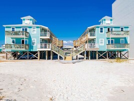 A beachfront property with two multi-story buildings featuring balconies and wooden decks, connected by a central staircase.