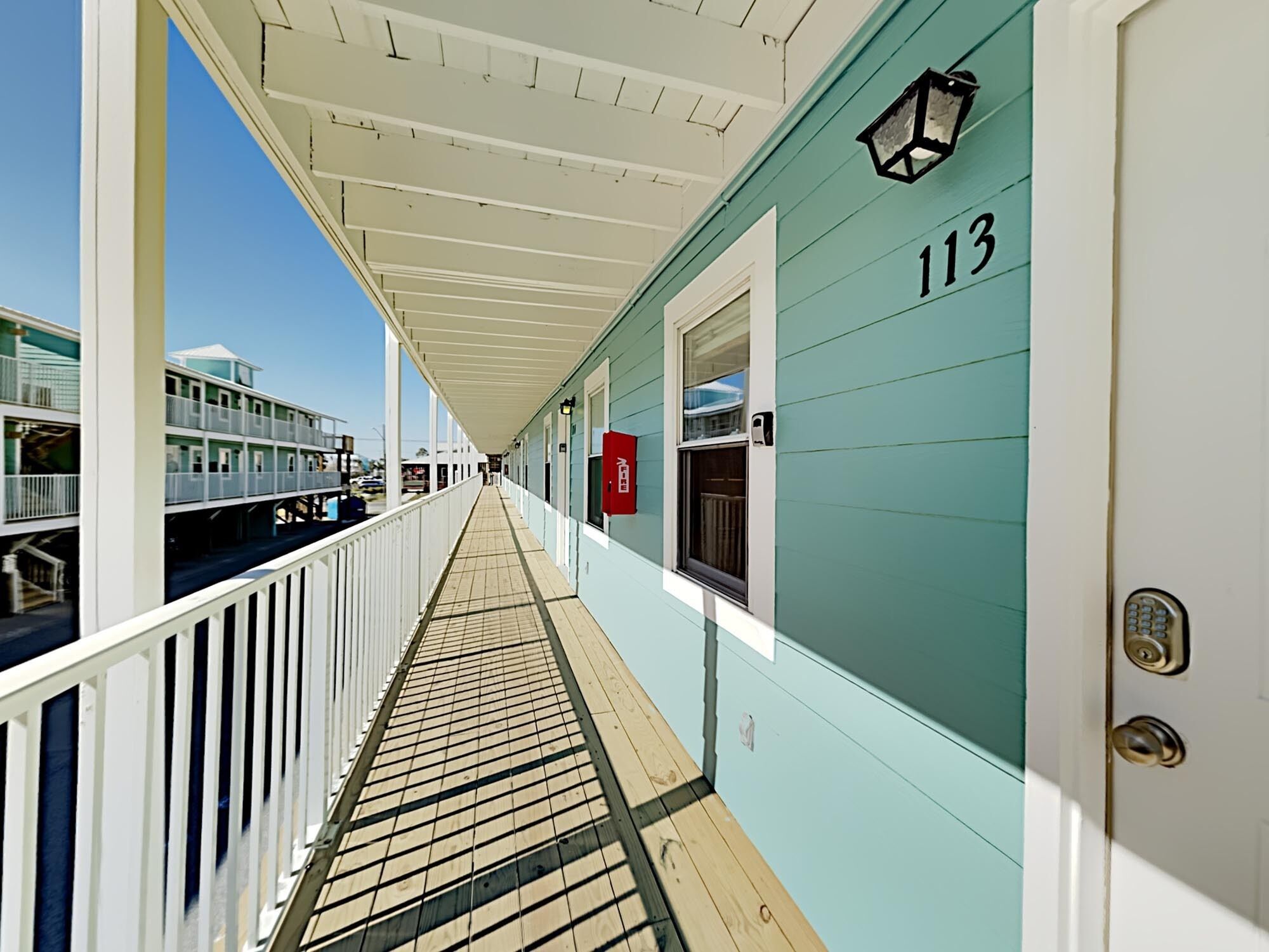 A corridor with a wooden deck, a white railing, and a teal-colored building with the number 113.