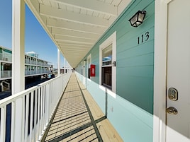A corridor with a wooden deck, a white railing, and a teal-colored building with the number 113.