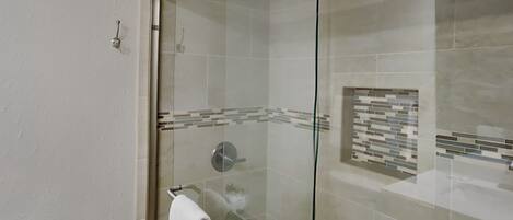 A glass-enclosed shower with a handrail, a tiled wall with a mosaic pattern, and a white towel hanging on a metal towel rack.