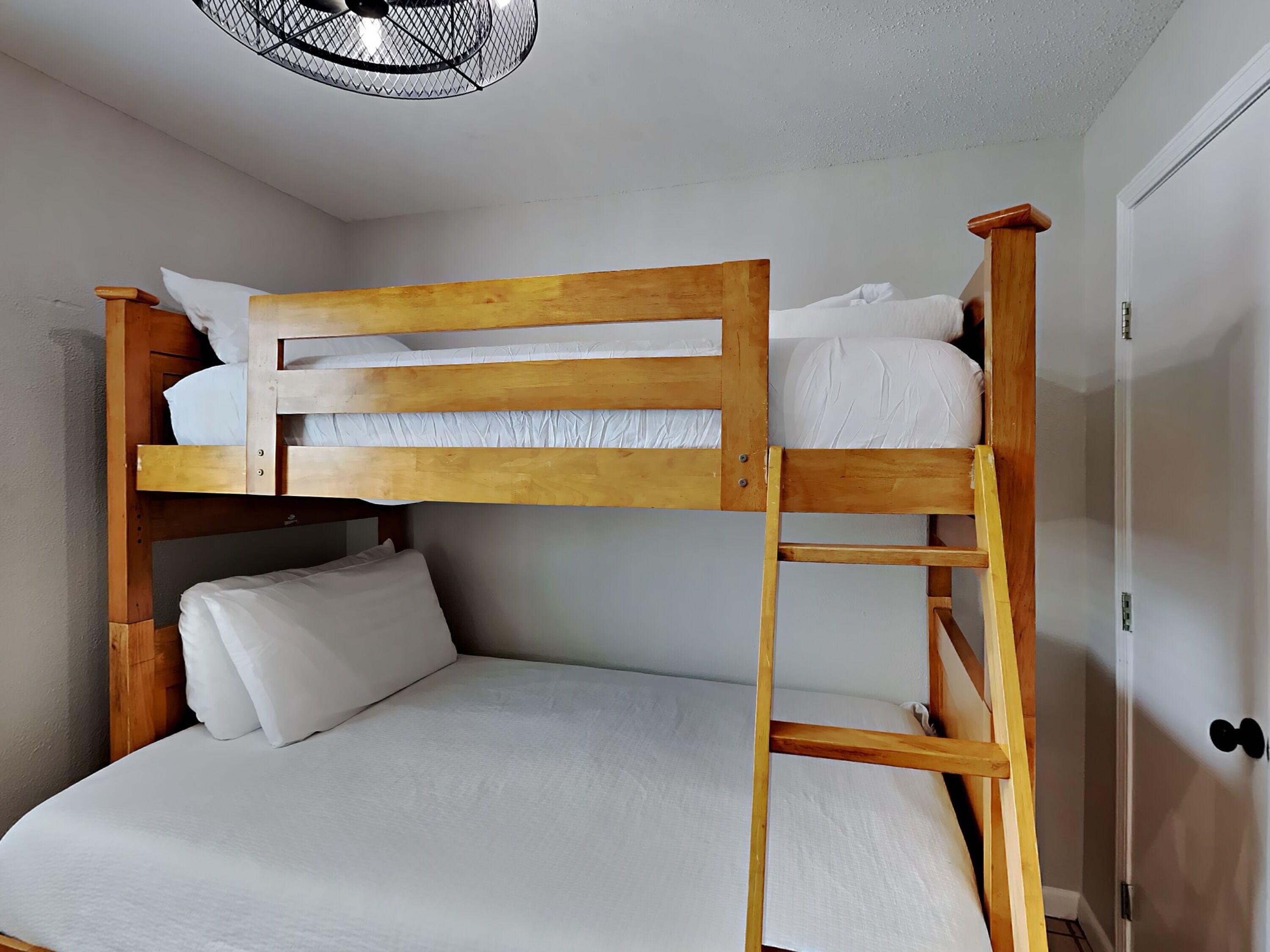 Un lit superposé avec une structure en bois et une literie blanche, une échelle et un ventilateur de plafond.