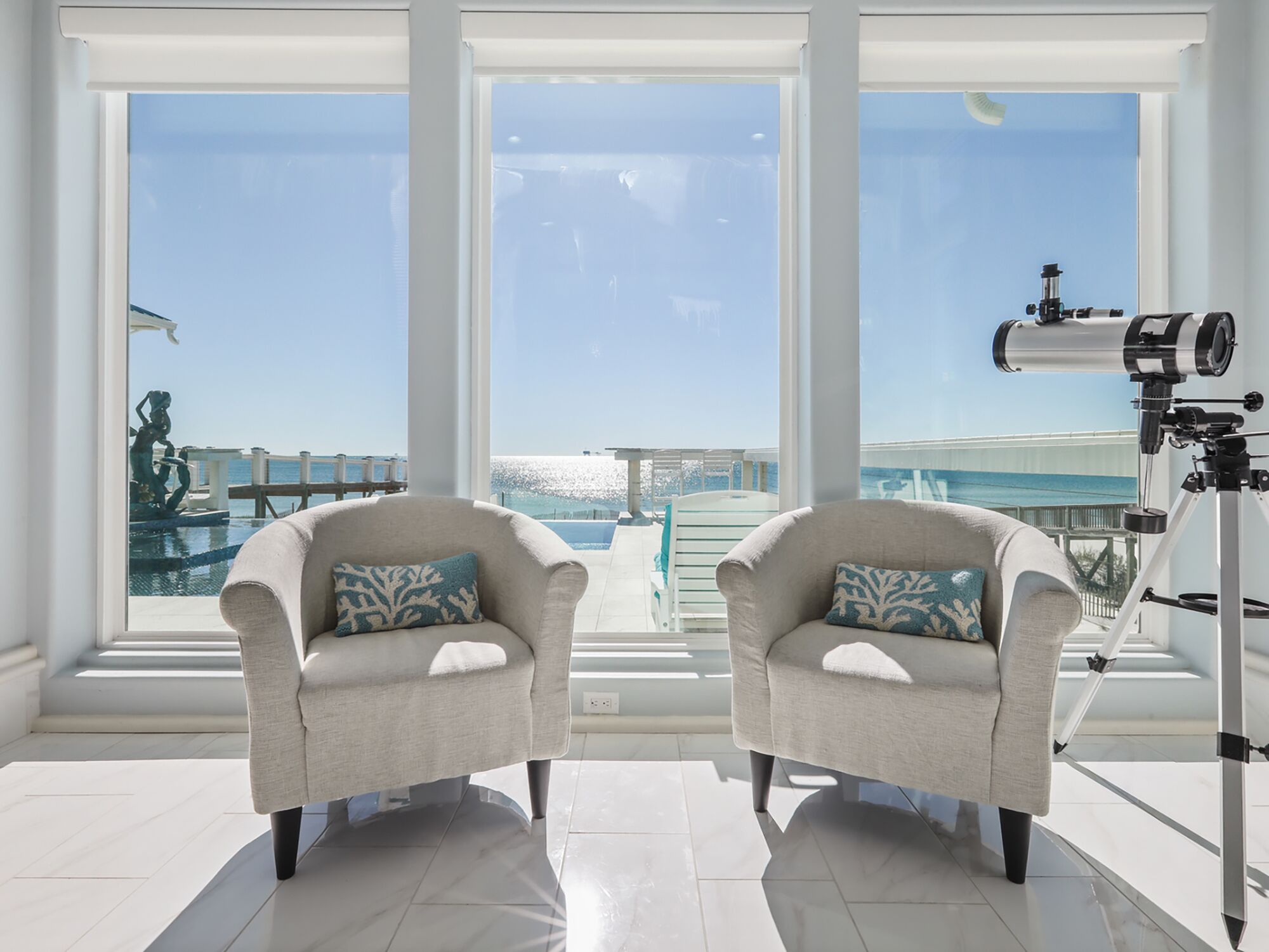 A room with two armchairs, a telescope, and a view of the ocean through large windows.