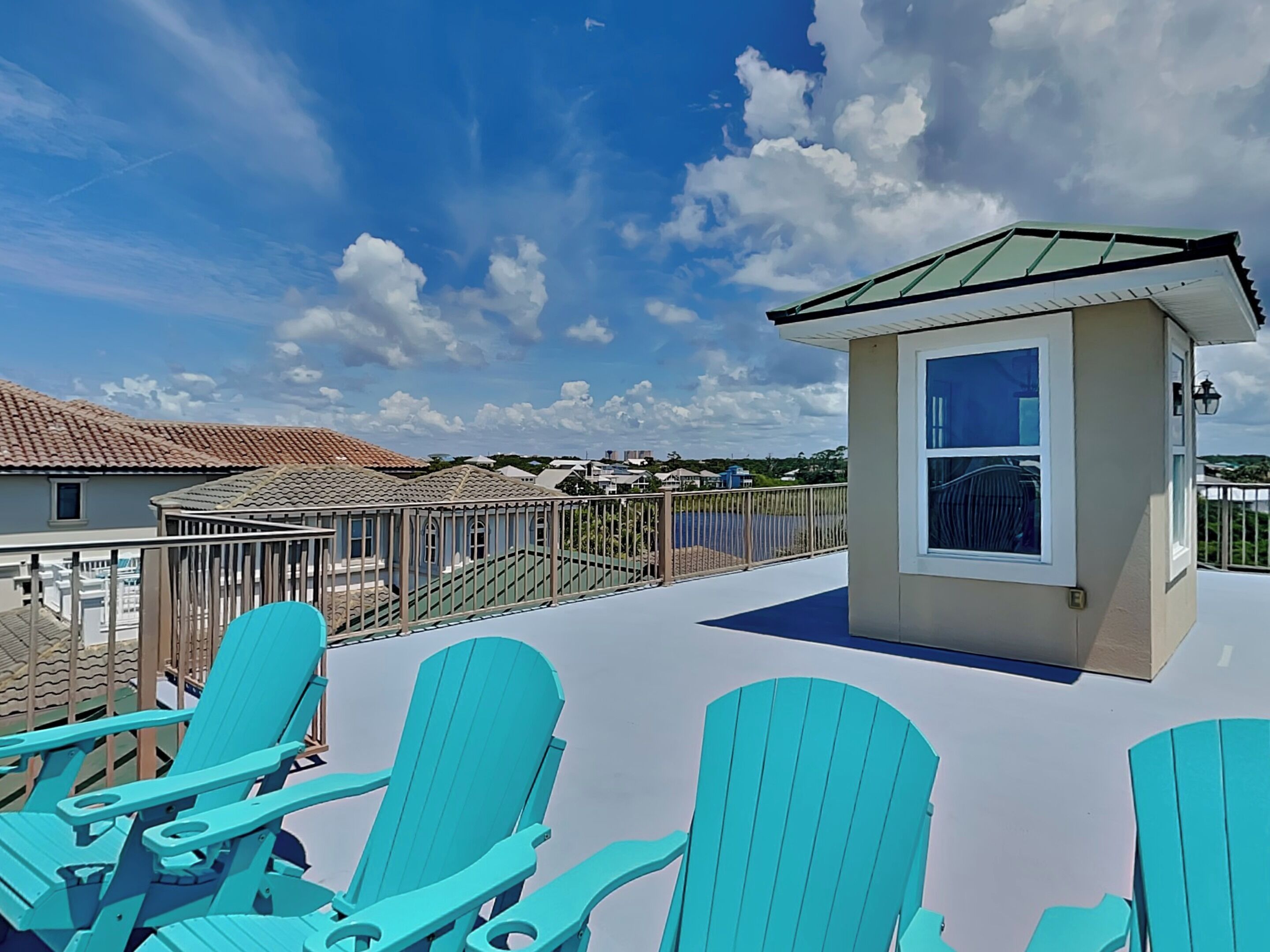 A rooftop terrace with turquoise Adirondack chairs, a small building with a green roof, and a view of neighboring houses.