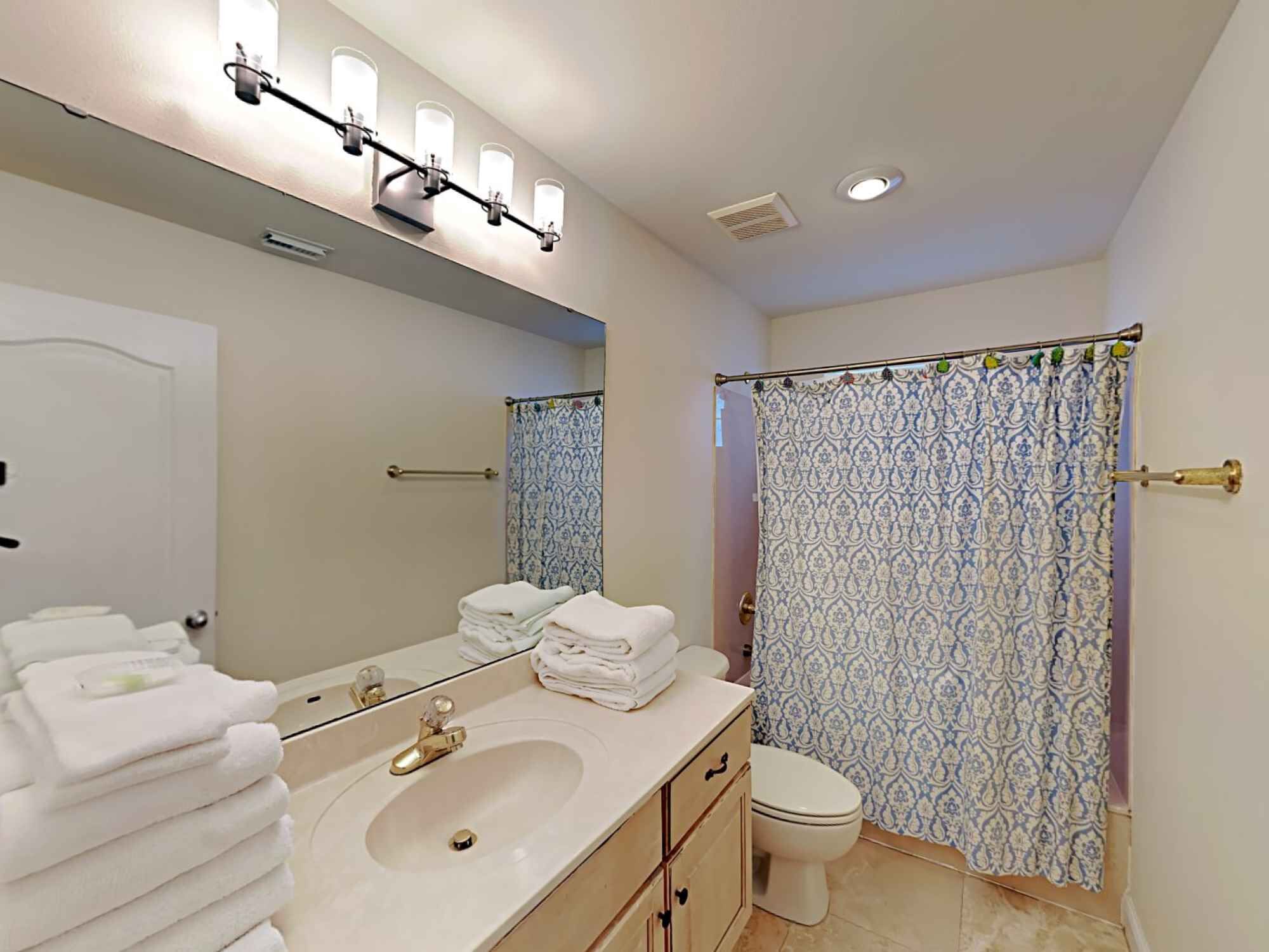 A bathroom with a sink, mirror, and toilet. There are towels stacked on the sink and a shower curtain with a pattern.