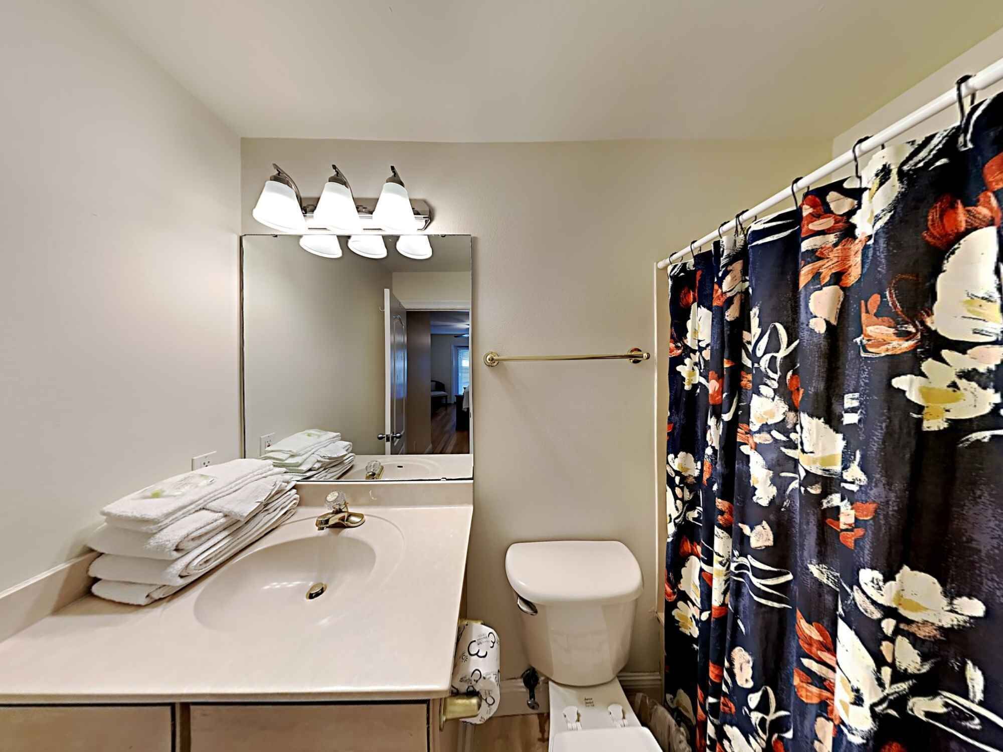 Een badkamer met een wit wastafel, toilet en een spiegel boven het wastafel. Er is een handdoekhouder met opgevouwen handdoeken en een douchegordijn met een bloemenpatroon.