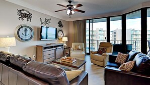 A modern living room with a leather sofa, a beige armchair, and a dark brown sofa. There is a flat-screen TV mounted on the wall, a ceiling fan, and a large window offering a view of the city. - Phoenix on the Bay by Southern Vacation Rentals (Orange Beach)