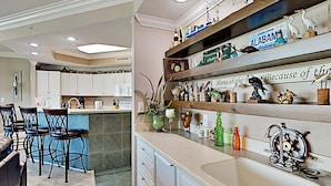A kitchen with white cabinets, a central island with bar stools, and decorative items on shelves. - Phoenix on the Bay by Southern Vacation Rentals (Orange Beach)