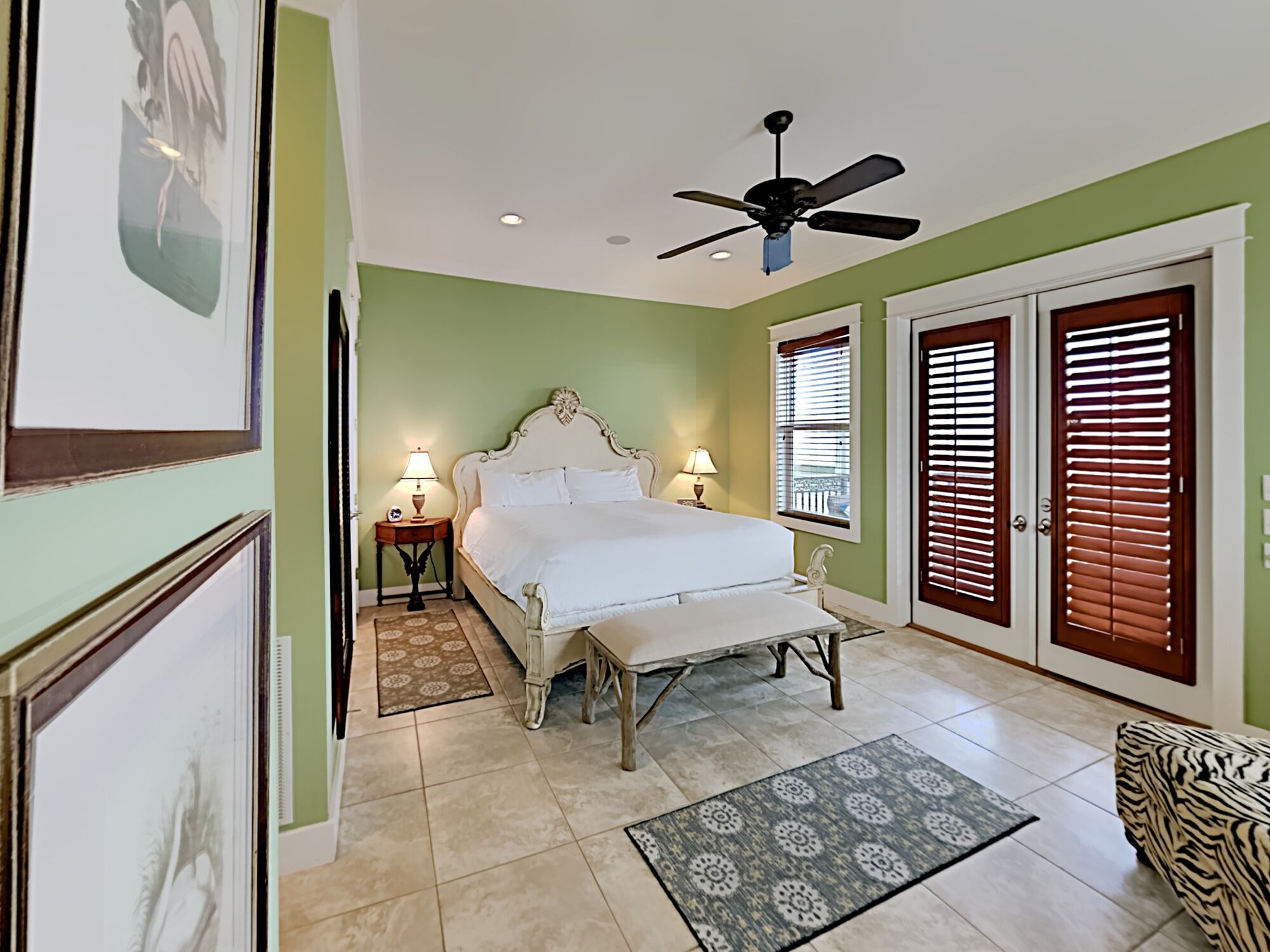A bedroom with a bed, two bedside tables with lamps, a ceiling fan, a window with blinds, and a door with a window.