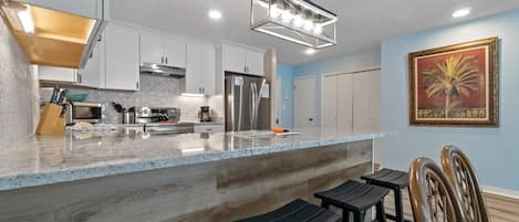 A modern kitchen with white cabinets, a stainless steel refrigerator, and a marble countertop. There are black stools and a wooden chair near the counter.