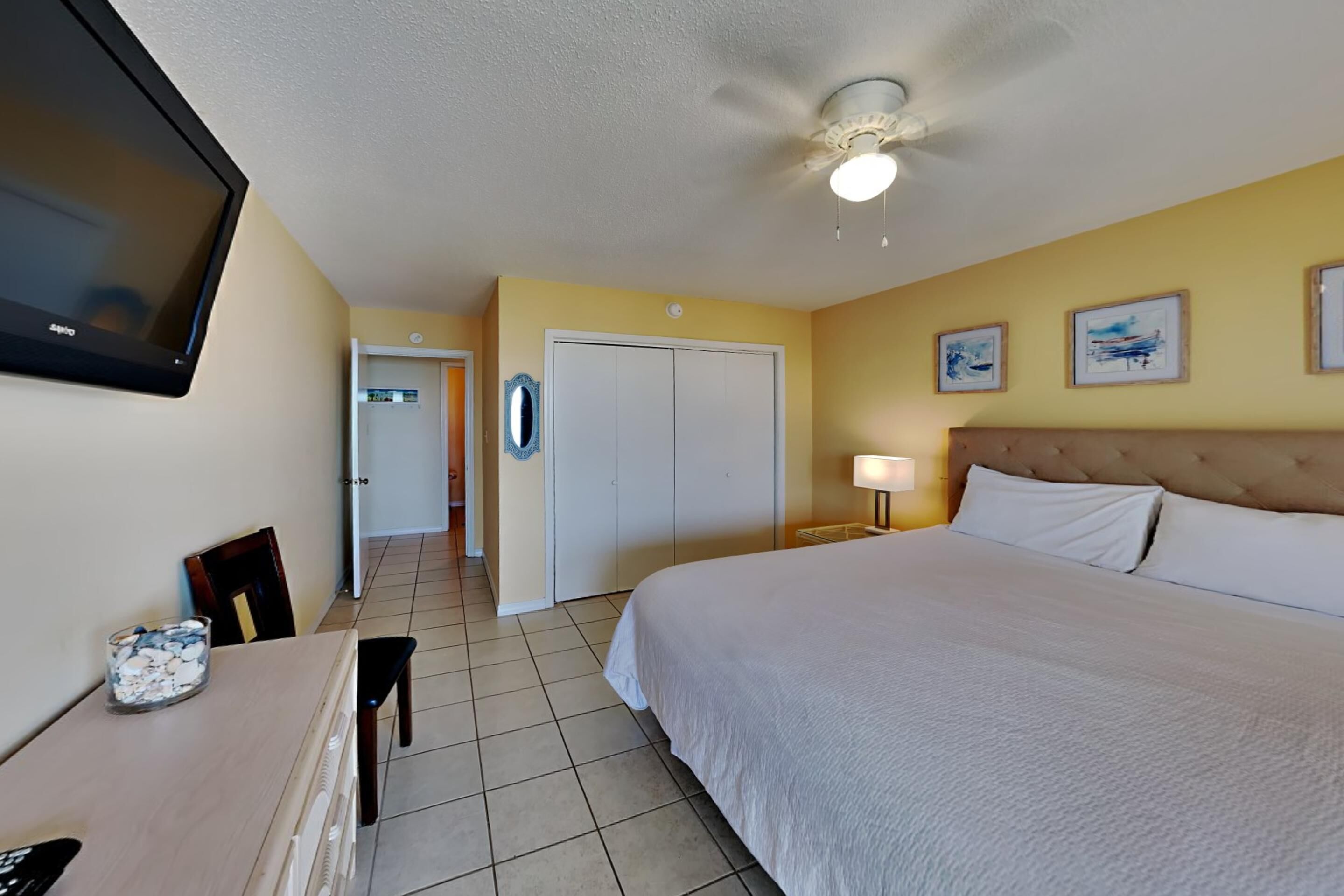 A hotel room with a bed, a desk with a lamp, a TV, and a door leading to another room.