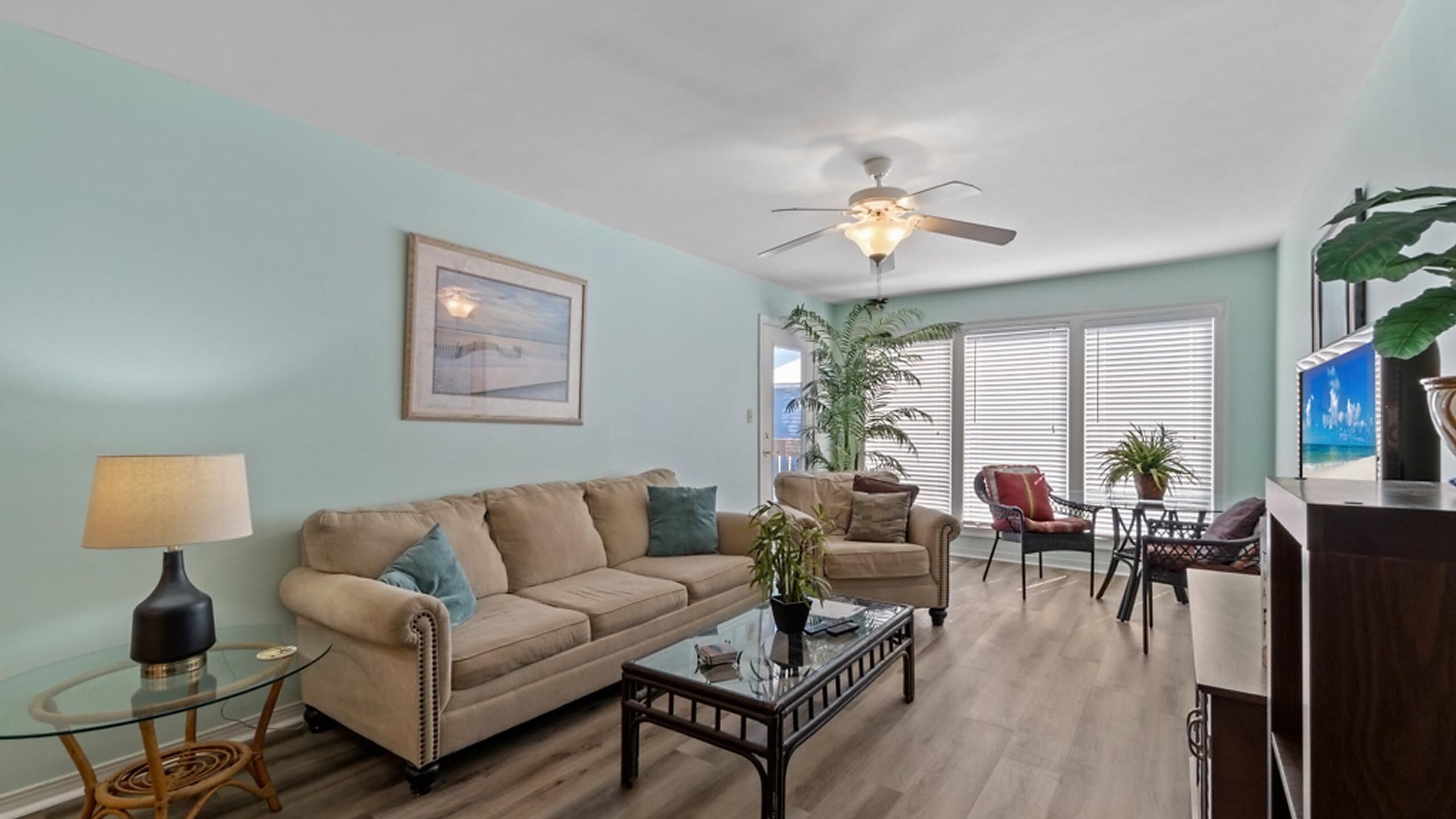 Un salon avec un canapé beige, une table basse en verre, un ventilateur de plafond et une grande fenêtre avec des stores.