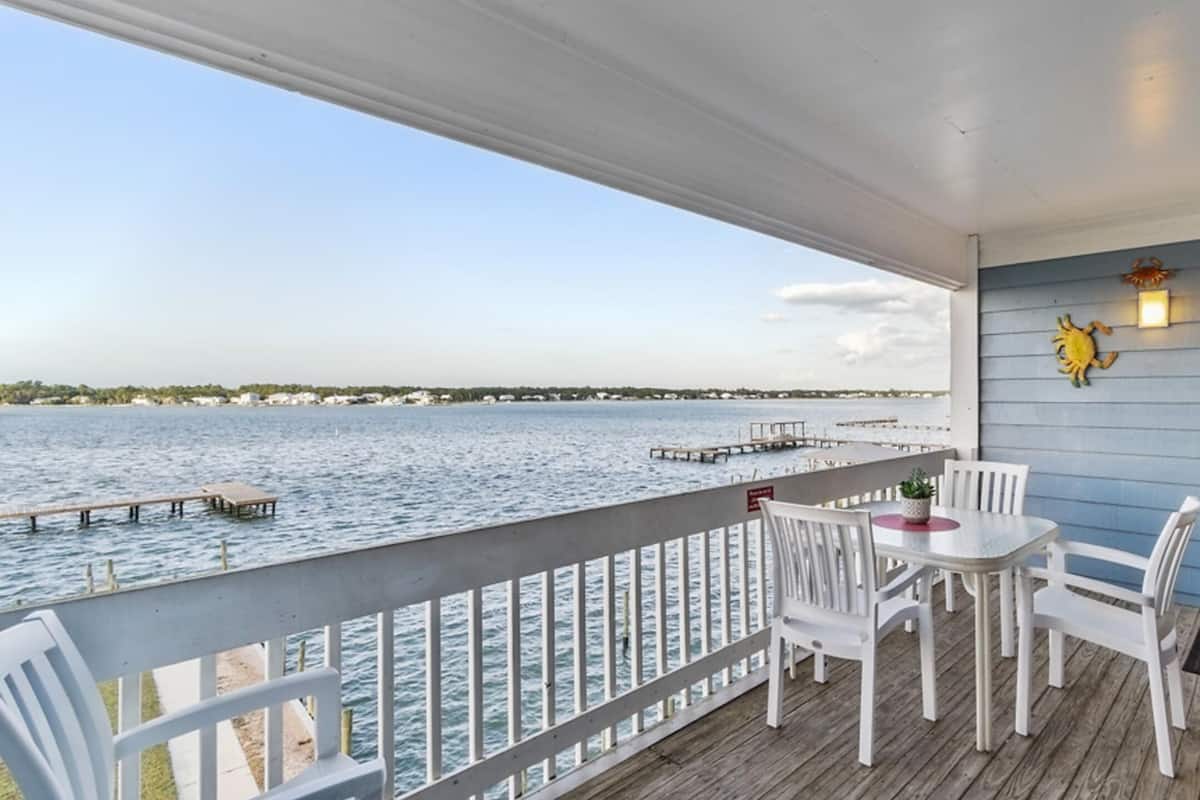 A covered outdoor deck with a view of the water, white chairs, a table, and decorative wall lights.