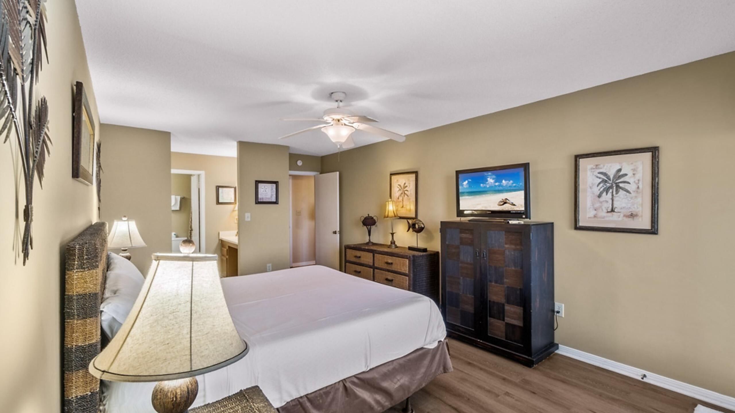 A hotel room with a bed, a ceiling fan, a television, a dresser, and wall art.