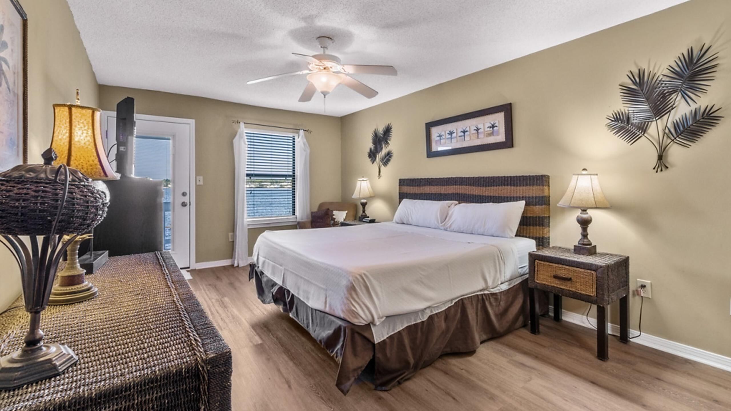 A bedroom with a bed, nightstands, a ceiling fan, a window with blinds, and a door.