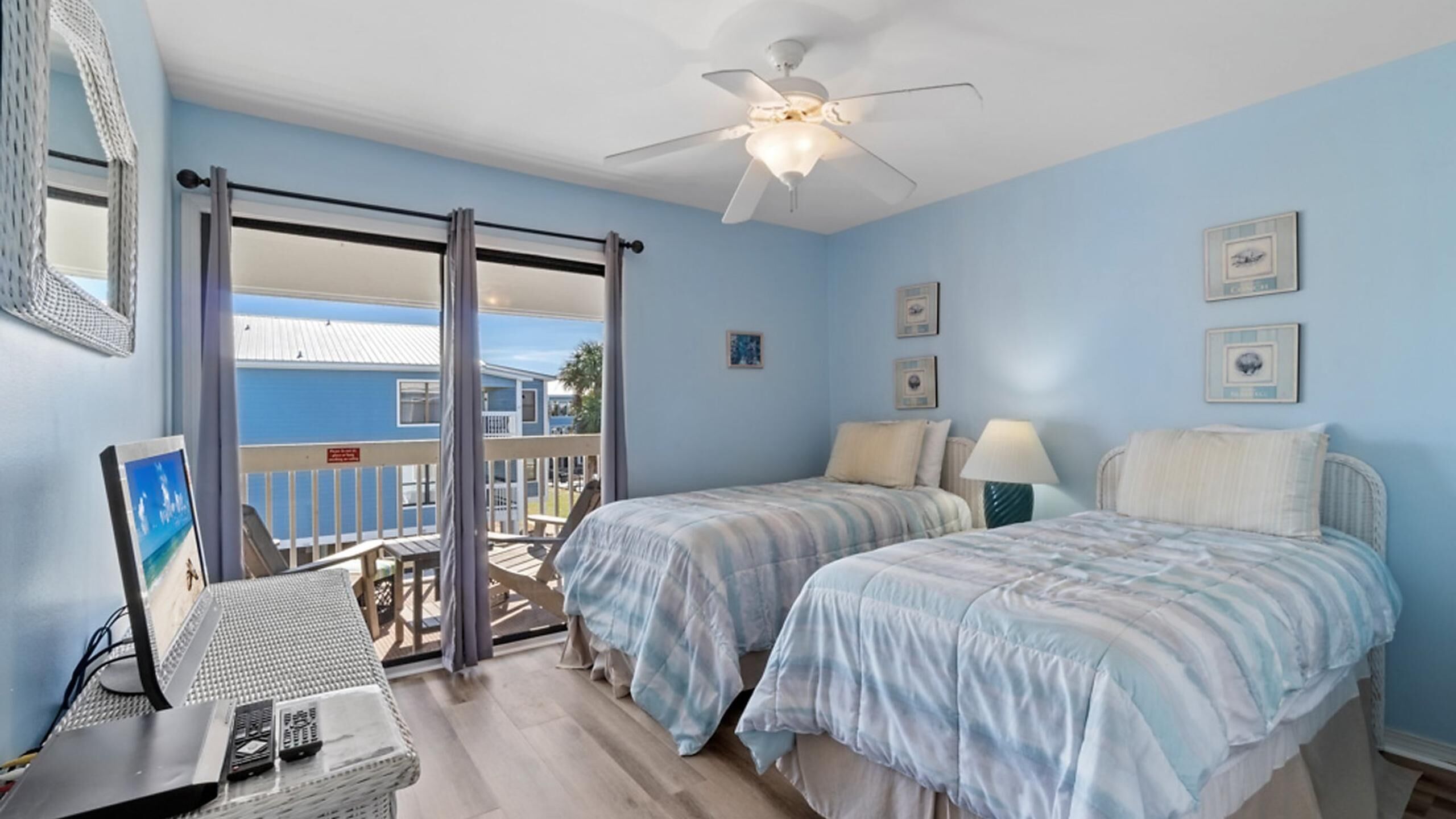 A room with two beds, a ceiling fan, and a view of a blue building through a sliding door.