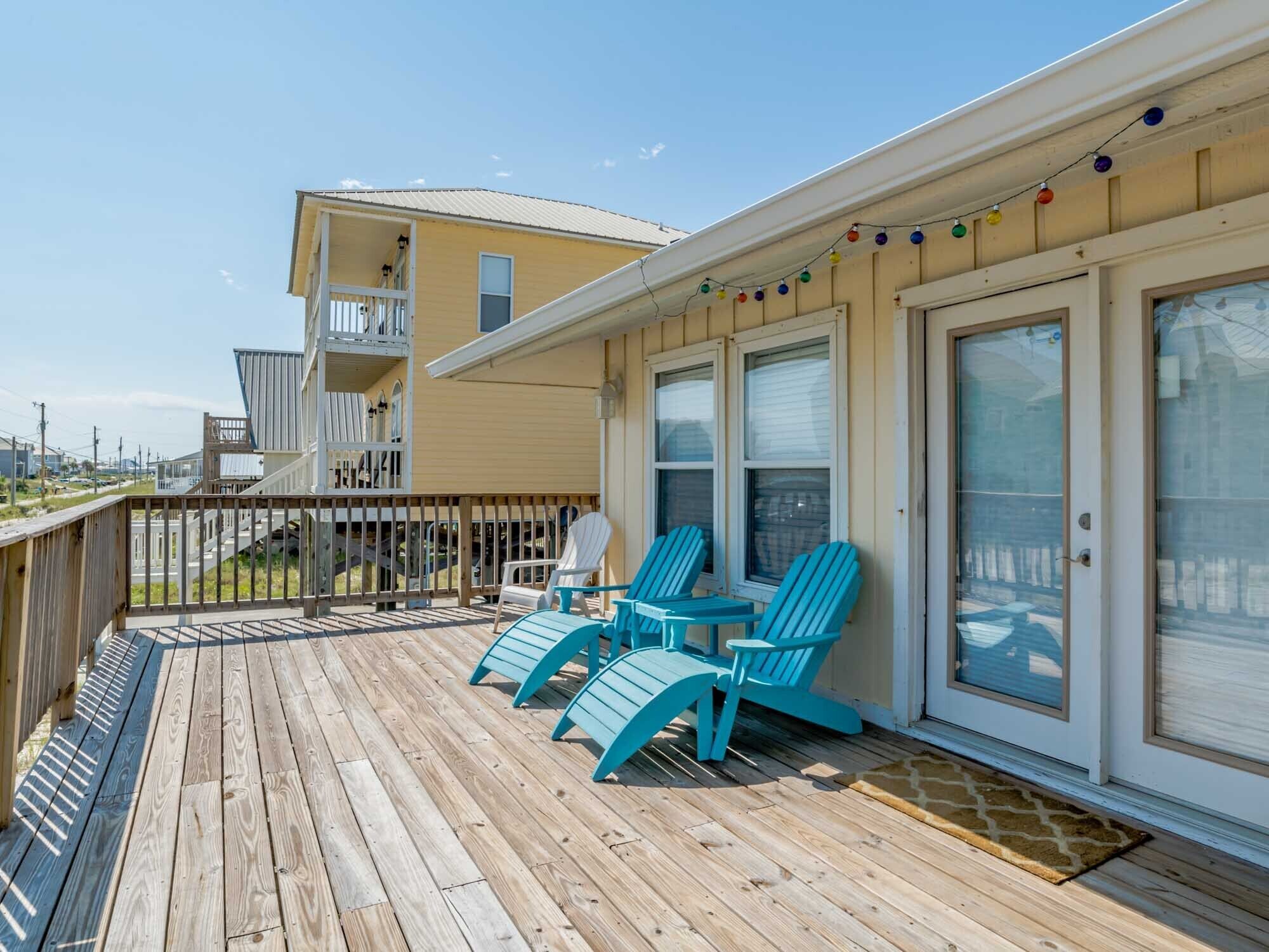 A wooden deck with turquoise chairs and a table, a balcony with a railing, and a building with large windows and a door.