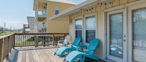 Une terrasse en bois avec des chaises turquoise et une table, un balcon doté d’une balustrade, et un bâtiment avec de grandes fenêtres et une porte.