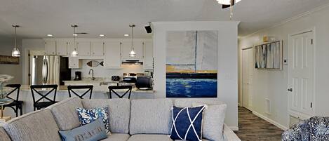 A modern living room with a sectional sofa, a dining area with a table and chairs, a kitchen with white cabinets, and a ceiling fan.