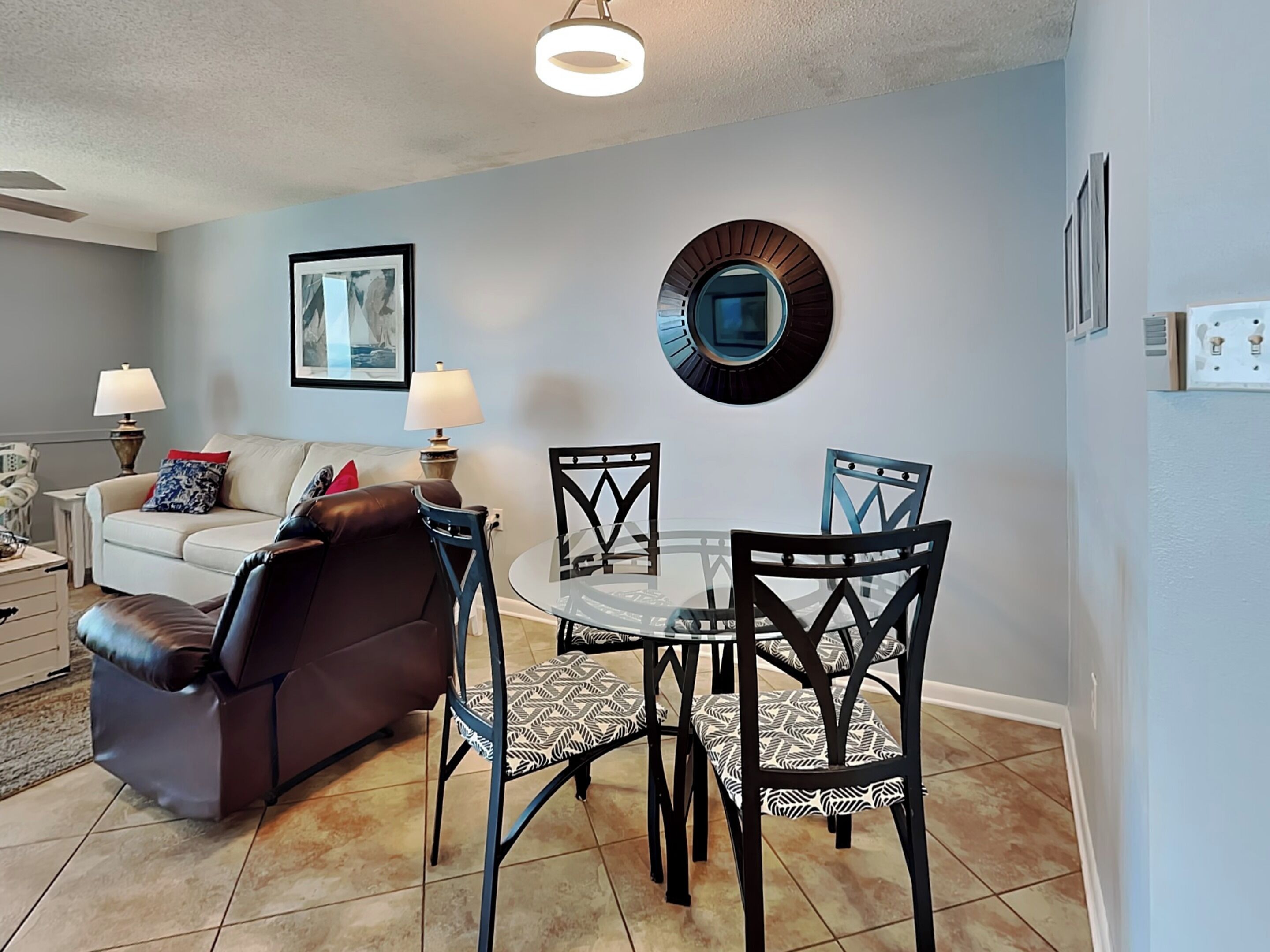 Un salon avec un canapé en cuir marron, une table basse en verre et des fauteuils à motifs. Un tableau encadré est accroché au mur et un miroir rond se trouve au-dessus de la table.