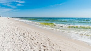 A wide sandy beach with gentle waves, clear skies, and distant buildings. - Phoenix III by Southern Vacation Rentals (Orange Beach)