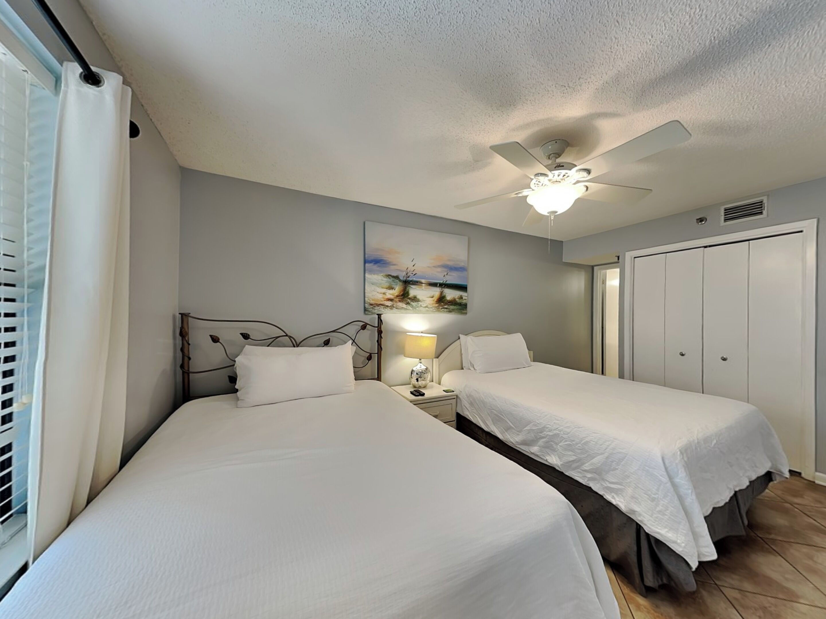 A hotel room with two beds, a ceiling fan, a nightstand with a lamp, a painting on the wall, and a wardrobe.