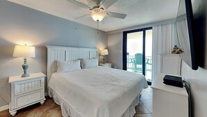 A bedroom with a bed, two bedside tables, a ceiling fan, a TV, and a balcony with chairs. - Phoenix III by Southern Vacation Rentals (Orange Beach)