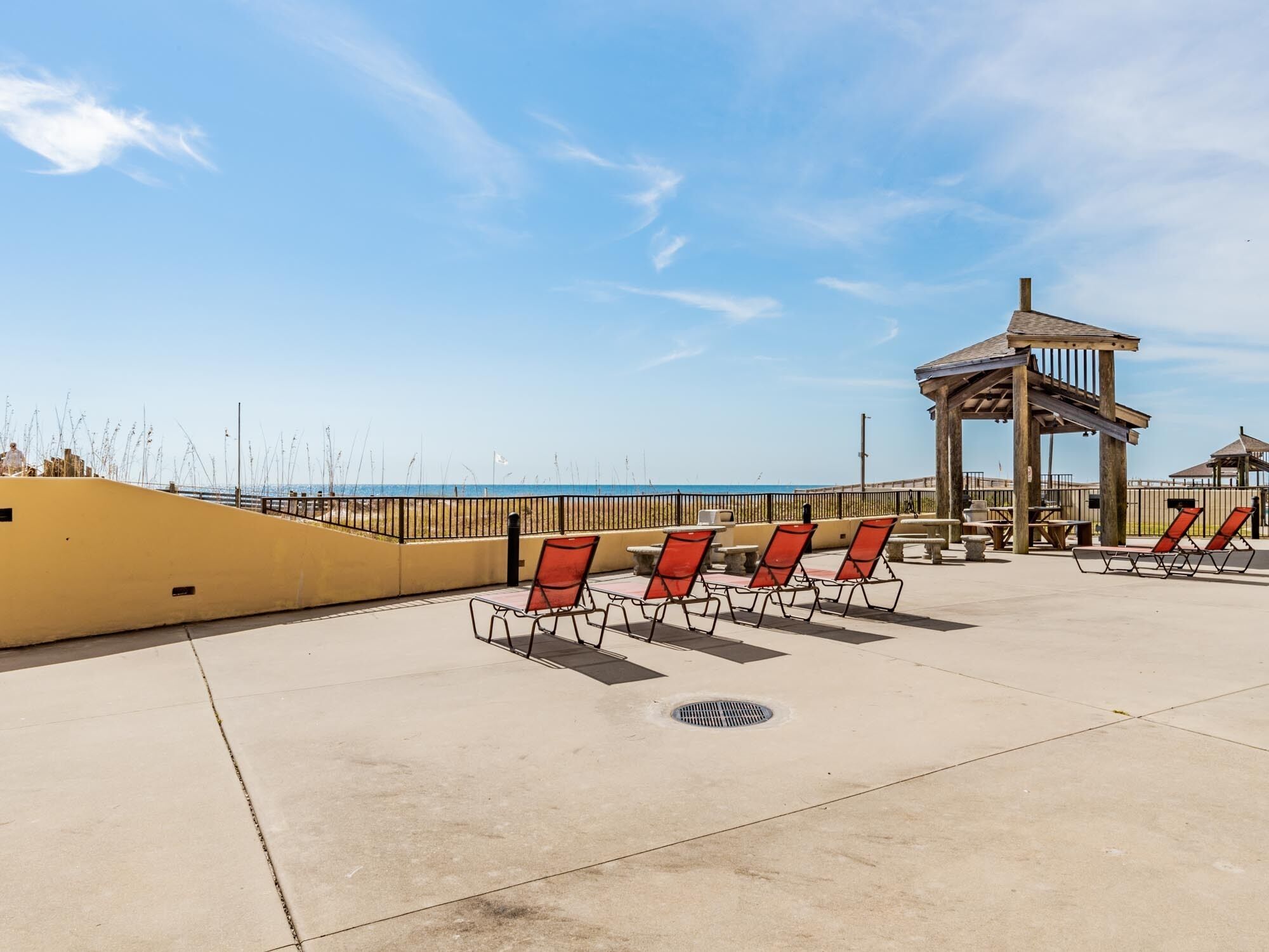 A beachside patio with sun loungers, a wooden gazebo, and a view of the ocean.