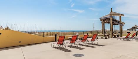 A beachside patio with sun loungers, a wooden gazebo, and a view of the ocean.