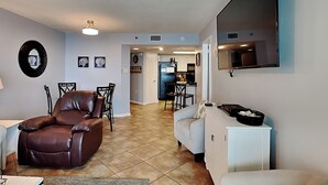 A living room with a brown leather sofa, a white armchair, and a dining area with a table and chairs. There is a kitchen in the background with a refrigerator and a coffee maker. - Phoenix III by Southern Vacation Rentals (Orange Beach)