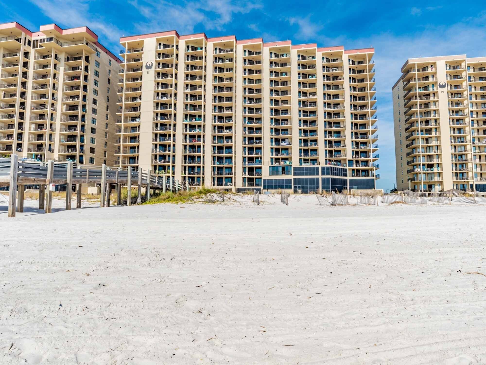 A beachfront high-rise hotel with multiple balconies, a sandy beach in the foreground, and a clear blue sky.