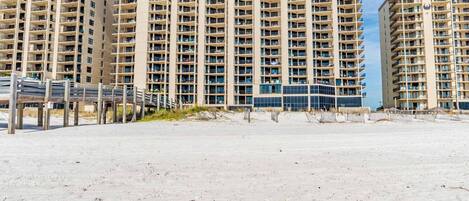 A beachfront high-rise hotel with multiple balconies, a sandy beach in the foreground, and a clear blue sky.