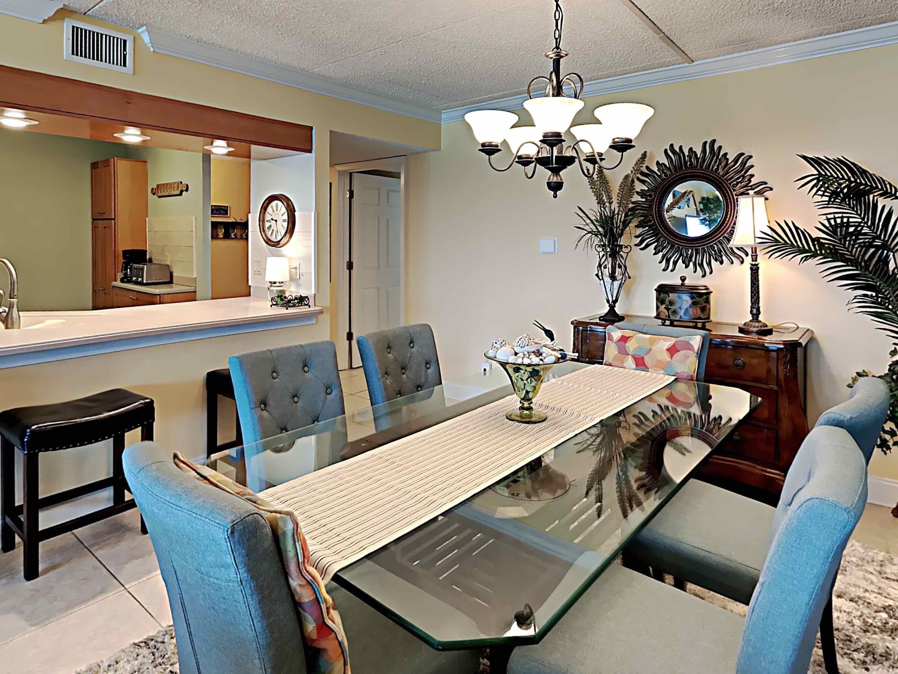 A dining area with a glass table, blue upholstered chairs, a chandelier, a mirror, and a decorative plant.