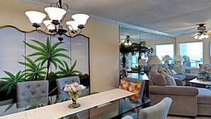 A modern living room with a glass dining table, grey upholstered chairs, a grey sofa, and a ceiling fan. - Island Echos #3K by Southern Vacation Rentals (Fort Walton Beach)