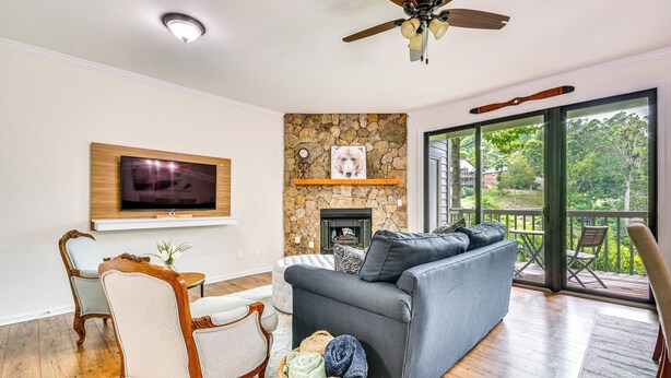 Een woonkamer met een stenen open haard, een grijze bank, houten stoelen, een televisie en een balkon met tuinmeubilair.