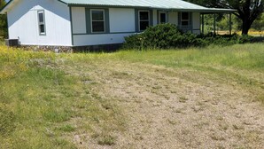 A single-story house with a green metal roof, a porch, and a large tree in the background. - Up Country Ranch House down a quiet lane (Mule Creek)