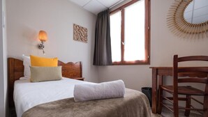 Un dormitorio con cama, una silla de madera, un escritorio, una ventana con cortinas, una lámpara de pared y un espejo decorativo.