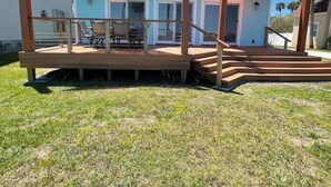 A wooden deck with a staircase leading to a covered area, a table and chairs, and a house in the background.